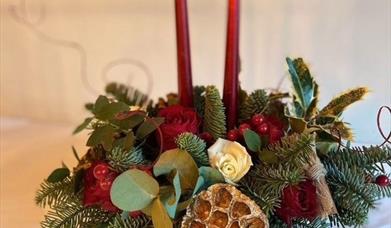 a Christmas table centrepiece decorated with holly, eucalyptus, roses and berries, with two red tapered candles coming out from the centre
