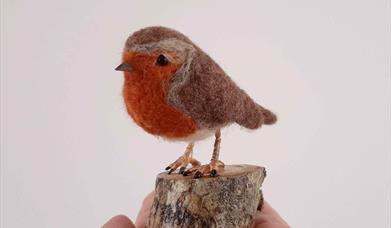 a person's hand holds a wool felted sculpture of a robin bird on a small articifical tree stump 