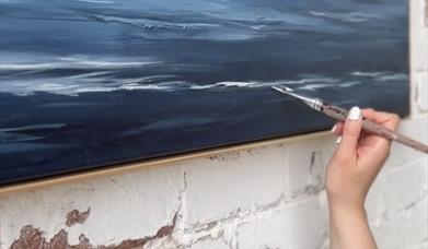 a hand holds a brush up to add finishing touches to a deep blue abstract painting of the sea