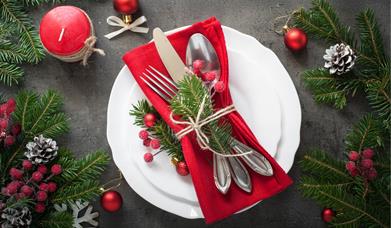 cutlery is wrapped together on a red napkin sitting on a plate, which is surrounded by festive decor including red berries, a red candle, pine cones a