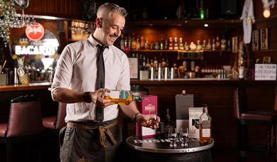 Bartender and mixologist, Ashely, pours whiskey in to a glass in front of a bar.
