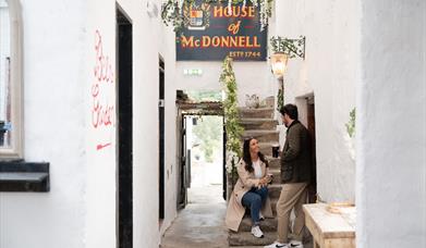 a woman sits on the steps at House of McDonnell while talking to a man who is standing, they are both holding drinks