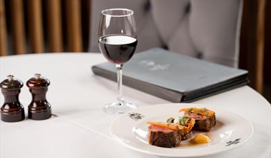 Image shows a chair and dining table covered in a white cloth, with a plate of food, a glass of red wine, a menu and salt/pepper grinders