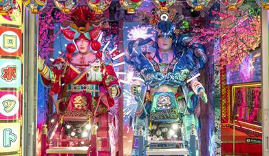 Two massive, highly stylized robotic figures with humanoid faces stand side-by-side in a vibrant, neon-lit window display. The figure on the left is a