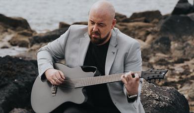 musician Jamie Sloan strums a guitar while sitting outside on coastal rocks