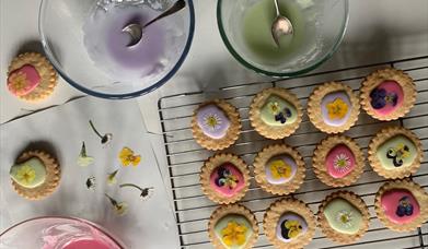 Image shows bowls of pastel coloured icing and some biscuits decorated with the iciing and with little edible flowers.