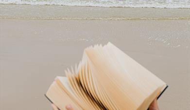 Close up image of hands holding a book with the sea and sand behind them,.
