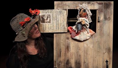 Person in a hat with red flowers on it standing next to a wooden door with a small hatch at the top through which a wooden puppet is sitting, hold a s