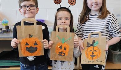 Imae of 3 children holding up their hand decorated pumpkin trick of treat bags. 