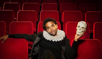 Hiran Abeysekera reclining across theatre seats holding a skull