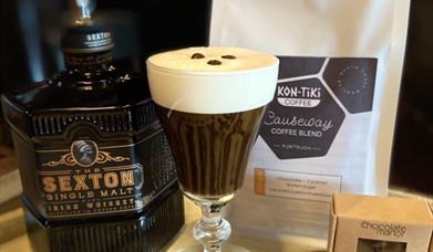 a bottle of Sexton Single Malt whiskey, an Irish Coffee with 3 coffee beans floating on the cream head, a bag of Kon-Tiki Causeway Coffee