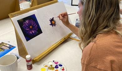 A woman painting a purple flower using a reference image attached to her canvas.