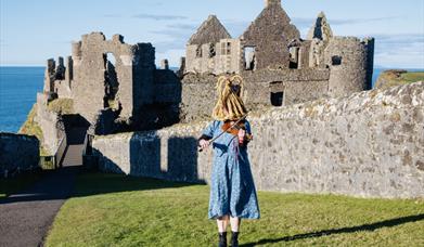 Person in a dress wearing a traditional 'mummers' mask plays the violin outside Dunluce Castle