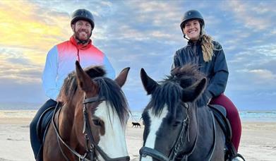 a man and woman sitting on horse back on the beach