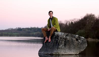 Image of Manchán Magan sitting on a rock in a lake.