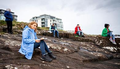 Participants of the workshop sit with sketch pads on coastal rocks in Portrush