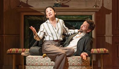 Image of a male and female character sitting closely together on a sofa, she is addressing the audience, he is looking at her.