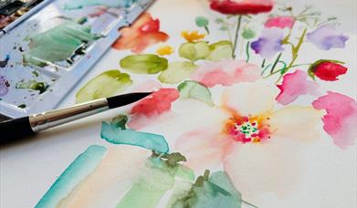 painting of flowers using watercolour paints rests beneath a small paintbrush and watercolour palette