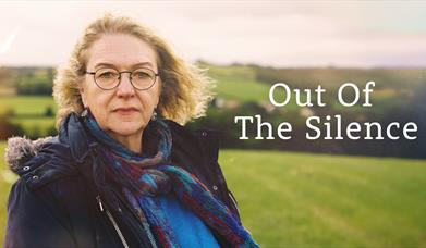 A woman with blond hair and round spectacles stands in a field looking towards the camera. Beside her the title "Out of the Silence" is written in whi
