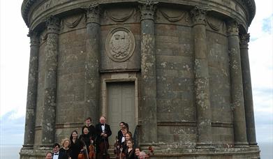 Donegal Chamber Orchestra pictured with their instruments standing on the steps of the Mussenden Temple