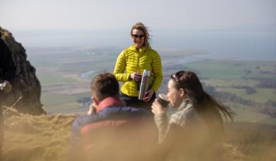 a man and woman sitting on a cliff drinking from cups. a woman stands in front of them with a thermal flask