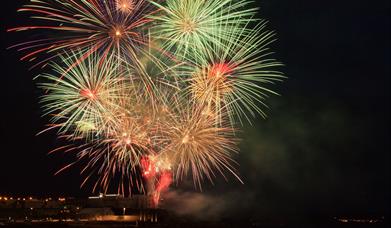 Fireworks burst to life above the sea and rocks, with the college in the background