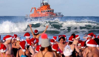 people in santa hats and swimwear watch on as a RNLI lifeboat cruises on the waves