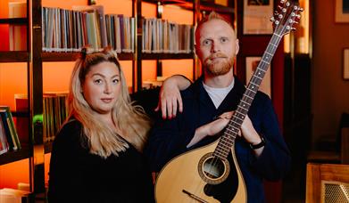 Image of Aideen McGinn and Martin Coyle tanding in from of a bookcase.  Martin is holding a guitar. 