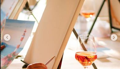 a person holds a pencil against a blank canvas resting on an easel. There is a glass of wine and a small plastic cup sitting on the table beside it.
