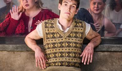 central characters of the play lined up behind a window all looking at the protagonist