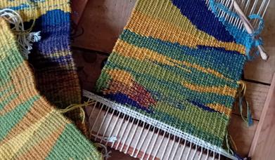 three colourful tapestries - one placed in a loom - sit on a wooden surface