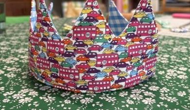 a birthday crown made of printed material that depicts cars and buses sits on a table covered with a green floral tablecloth
