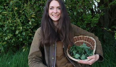 A woman stands smiling in the forest with a wicker basket full of foraged plants.