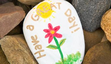 a picture of some brown stones with a white stone in the middle which has been painted to say 'Have a  Great Day' with a red flower and the sun