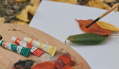 an artist's palette holding tubes of green, orange and yellow paint and a paintbrush sits on the forest floor beside a blank page which has leaves on 