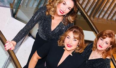 the three members of the band Leading Ladies stand dressed in glitzy black outfits on a staircase