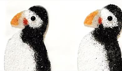 Two glass puffins facing left. They are flat against a white background. 