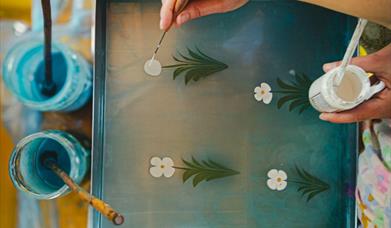 peoples' hands hold a paint brush to paint flowers on a painted flower stalk 