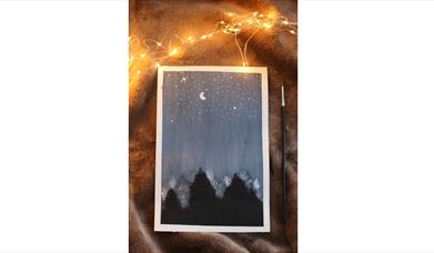 a painting of a nighttime landscape featuring shadowy trees and the moon and the stars rests on a faux fur blanket with fairy lights strewn on top