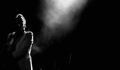 black and white image of male figure in shadows with a spotlight shining down on him.