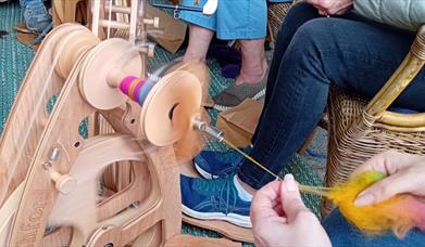 a group of people are seated and in the foreground, a person uses their hands to feed wool through a traditional wool spinning wheel