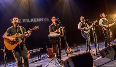 Image shows four men on stage playing instruments and singing into microphones
