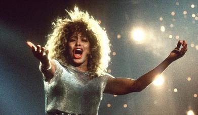 Tina Turner singing against sparkling lights with arms outstretched