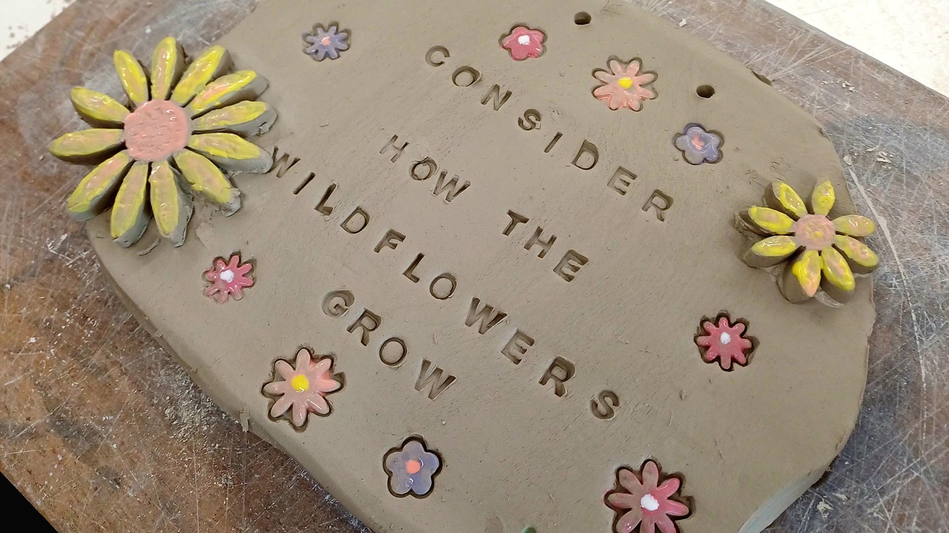 A ceramic sign with flowers carved into and embellishing it. It reads "Consider how the Wild Flowers grow"