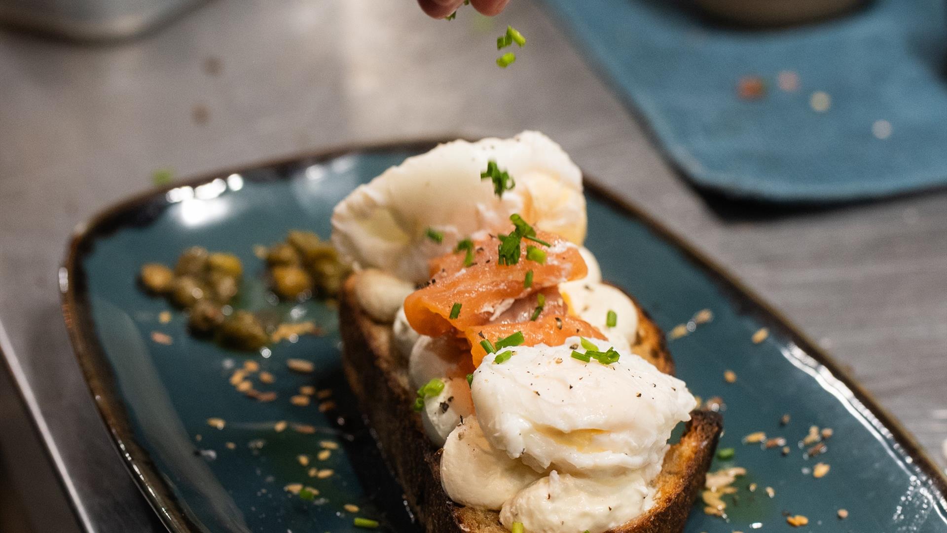 a persons hand sprinkles a herb garnish over poached eggs and salmon on sourdough bread