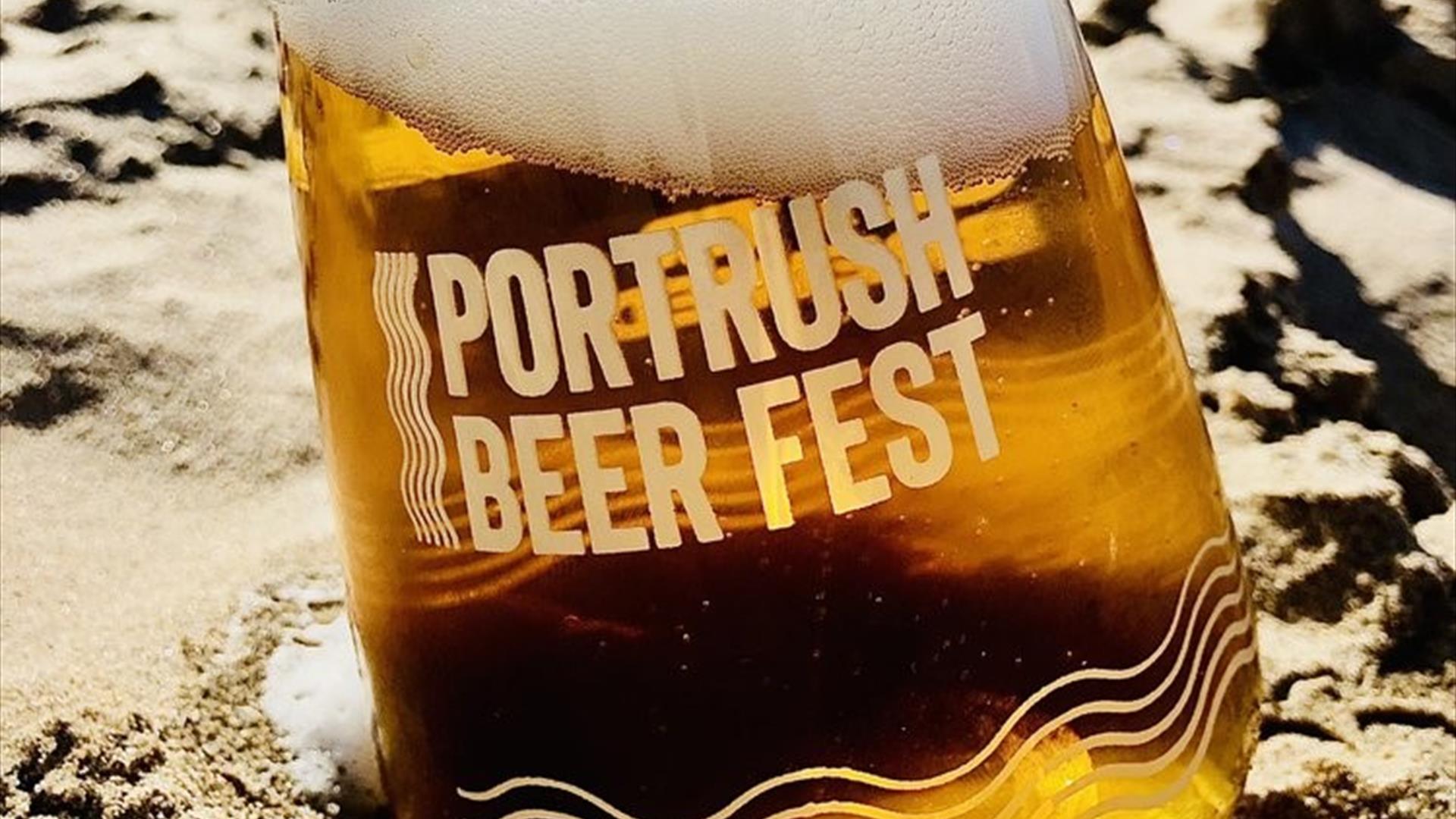 A glass of frothing beer on the sand. Text on the glass reads "Portrush Beer Fest"