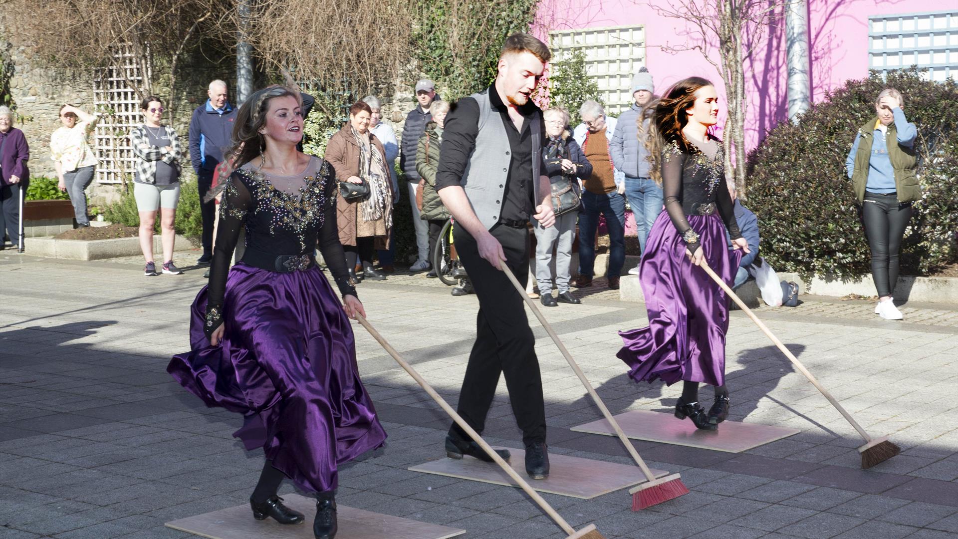 Irish dancers entertaining the crowds in Drumceatt Square, dressed in purple and black dresses with brooms in hand.