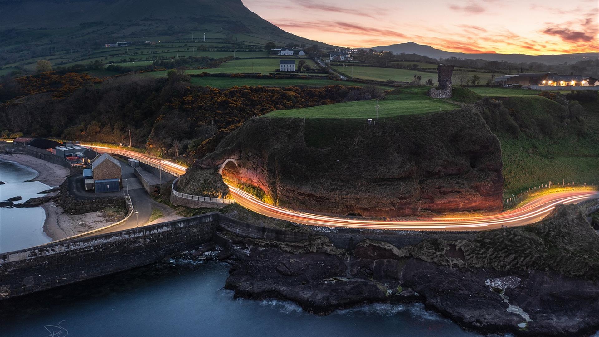 Panoramic view of Red Bay in Cushendall, County Antrim, with colourful boats docked in the small harbour and green hills in the background.