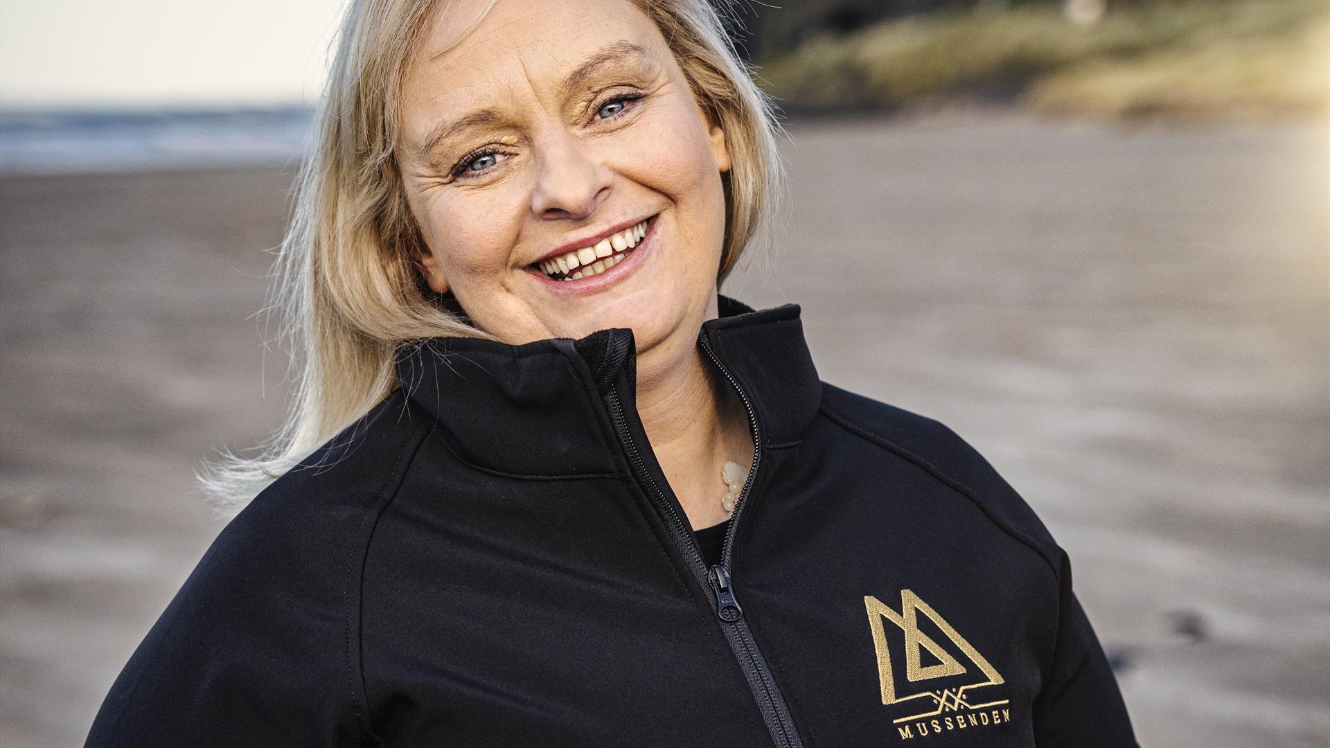 Claire O'Kane of Mussenden Sea Salt stands smiling beneath Mussenden Temple, wearing a black zip up fleece with her company logo in gold.
