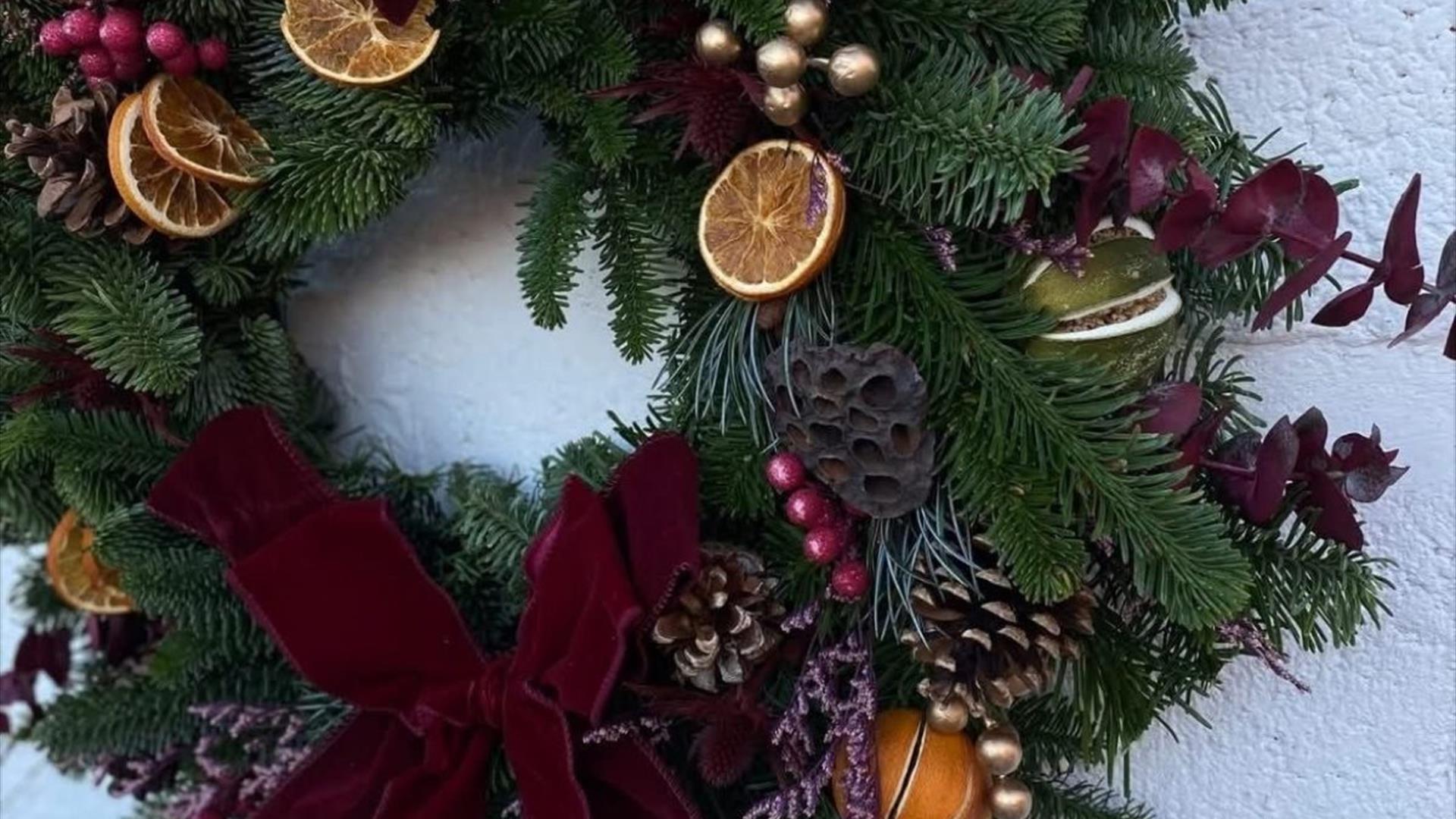a christmas wreath hangs on a wall and is decorated with a dark red velvet bow, dried oranges and red eucalyptus stems